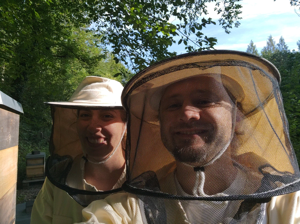 Zwei lächelnde Menschen in Imkerschleiern am Bienenstand, im Hintergrund Bäume und Bienenkästen